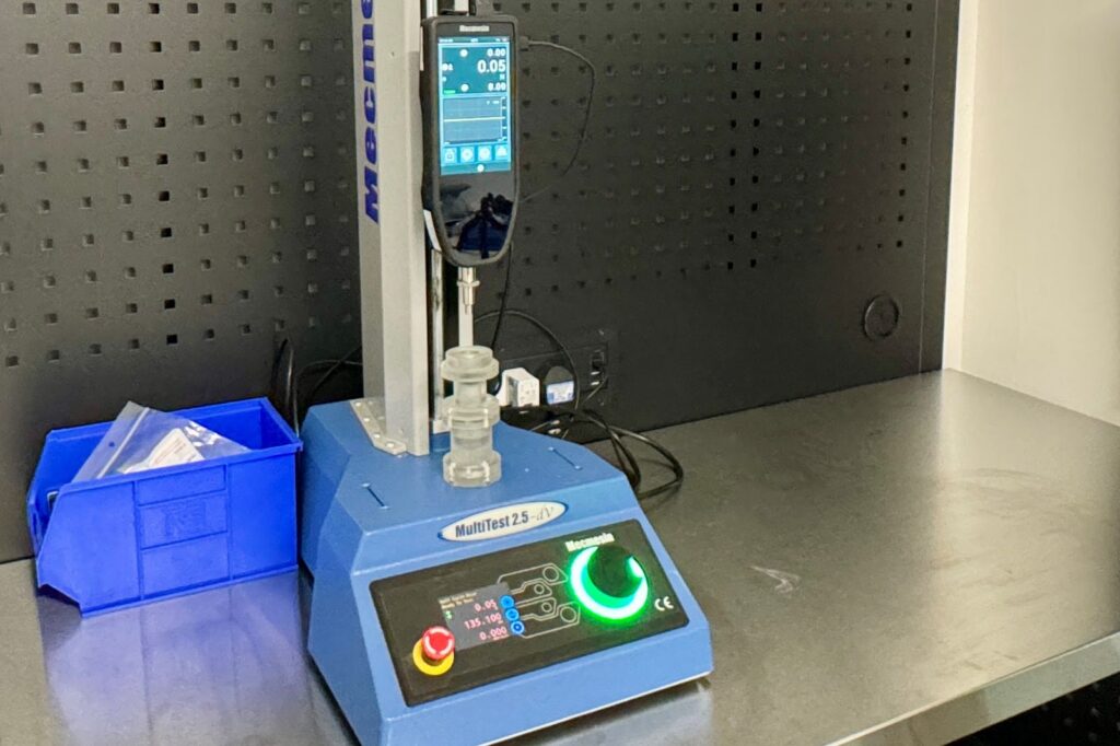 Mecmesin MultiTest 2.5-dV motorised force tester on the bench in the MDIN workshop, with a device component in the test fixture